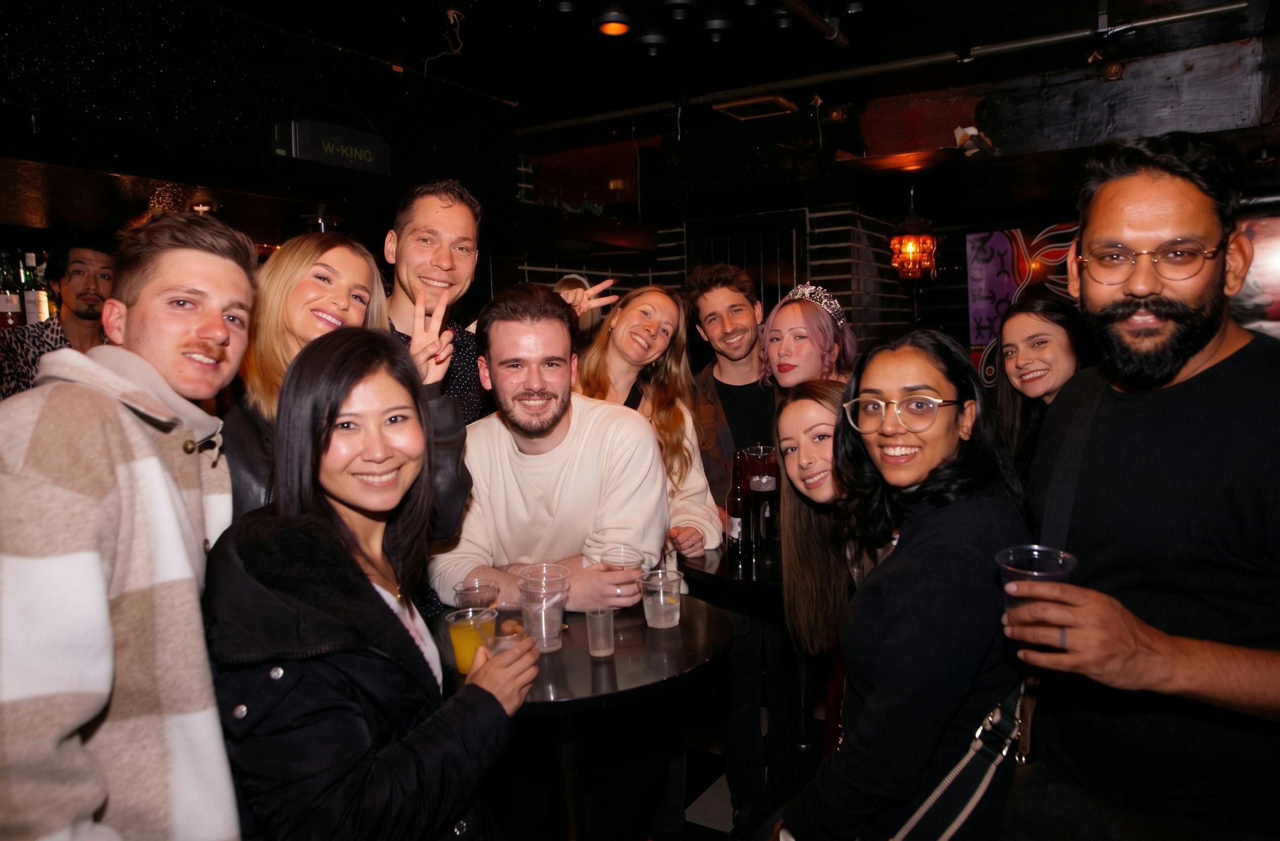 A group of people smiling and posing together in a dimly lit indoor setting, with drinks on a table in the foreground.