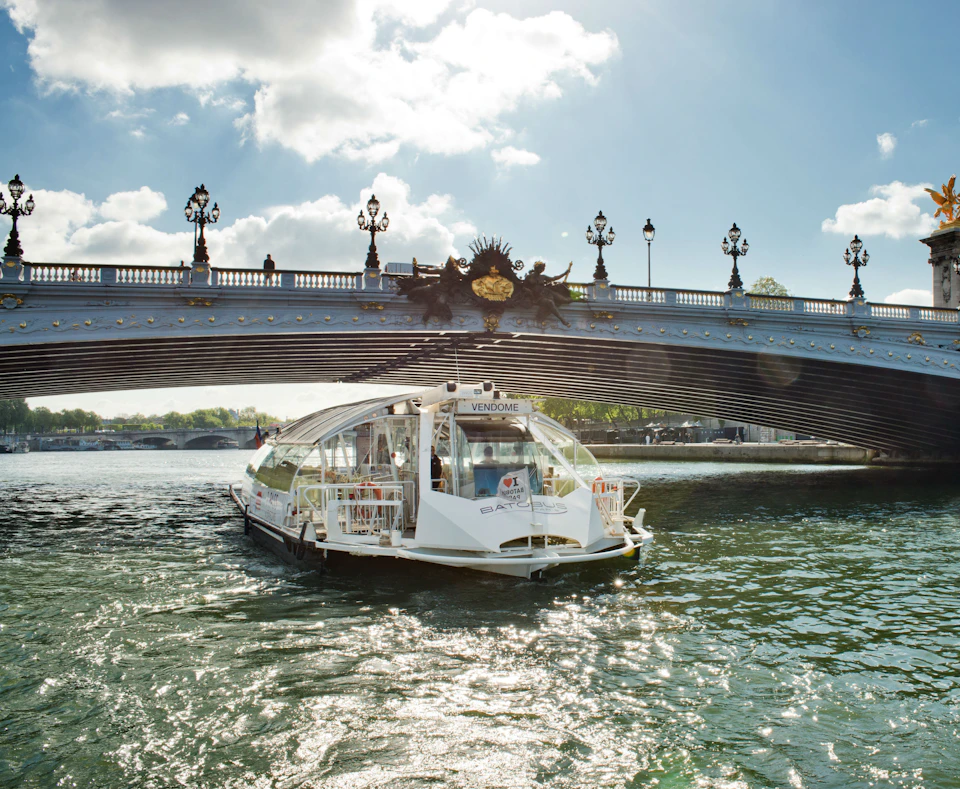 A boat with a transparent roof labeled "Vendome" passes under a decorated bridge on a sunny day.