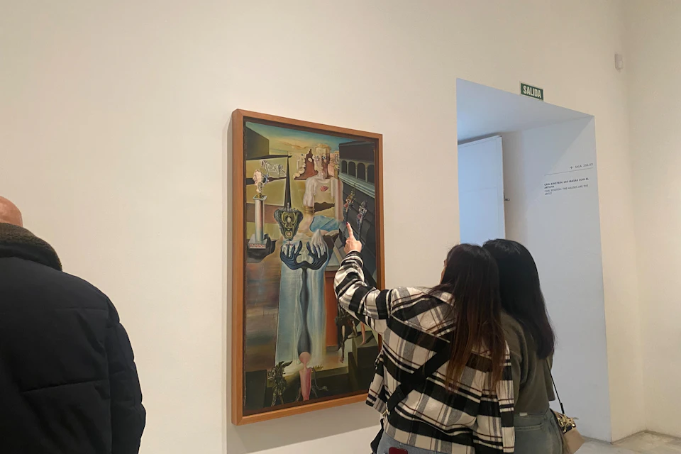 Two individuals observing and pointing at a surrealist painting in a gallery with a white wall and exit sign visible.