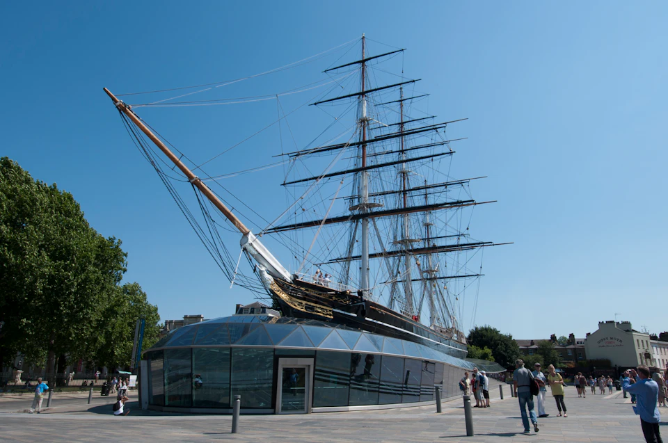 A tall ship with multiple masts is displayed above a modern glass structure. People are walking around the exhibit on a clear day.