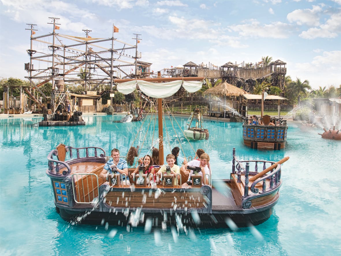 水しぶきとロープコースを背景に、遊園地で海賊をテーマにした水上ボートに乗る人々。