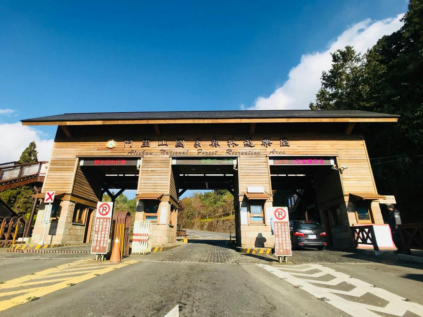 Entrance gate to a forest recreation area with a wooden structure, signs, and a car passing through on a sunny day.