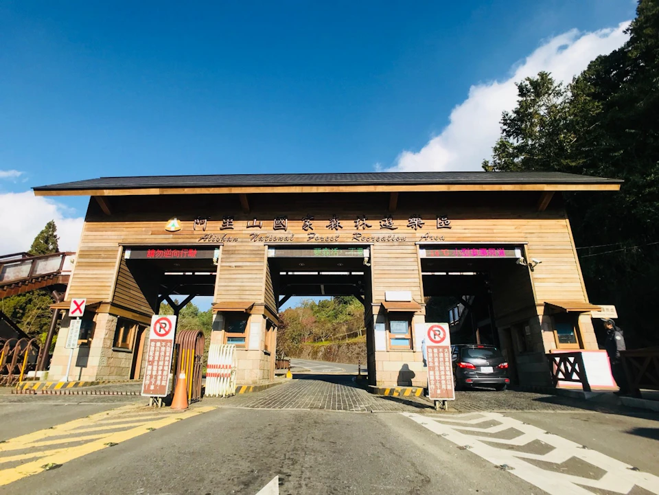 Entrance gate to a forest recreation area with wooden structure, signage, and cars on a sunny day.