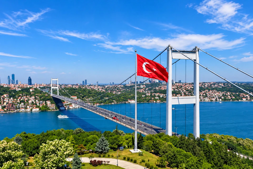 A scenic view of a suspension bridge over a body of water, with a Turkish flag in the foreground and cityscape in the background.
