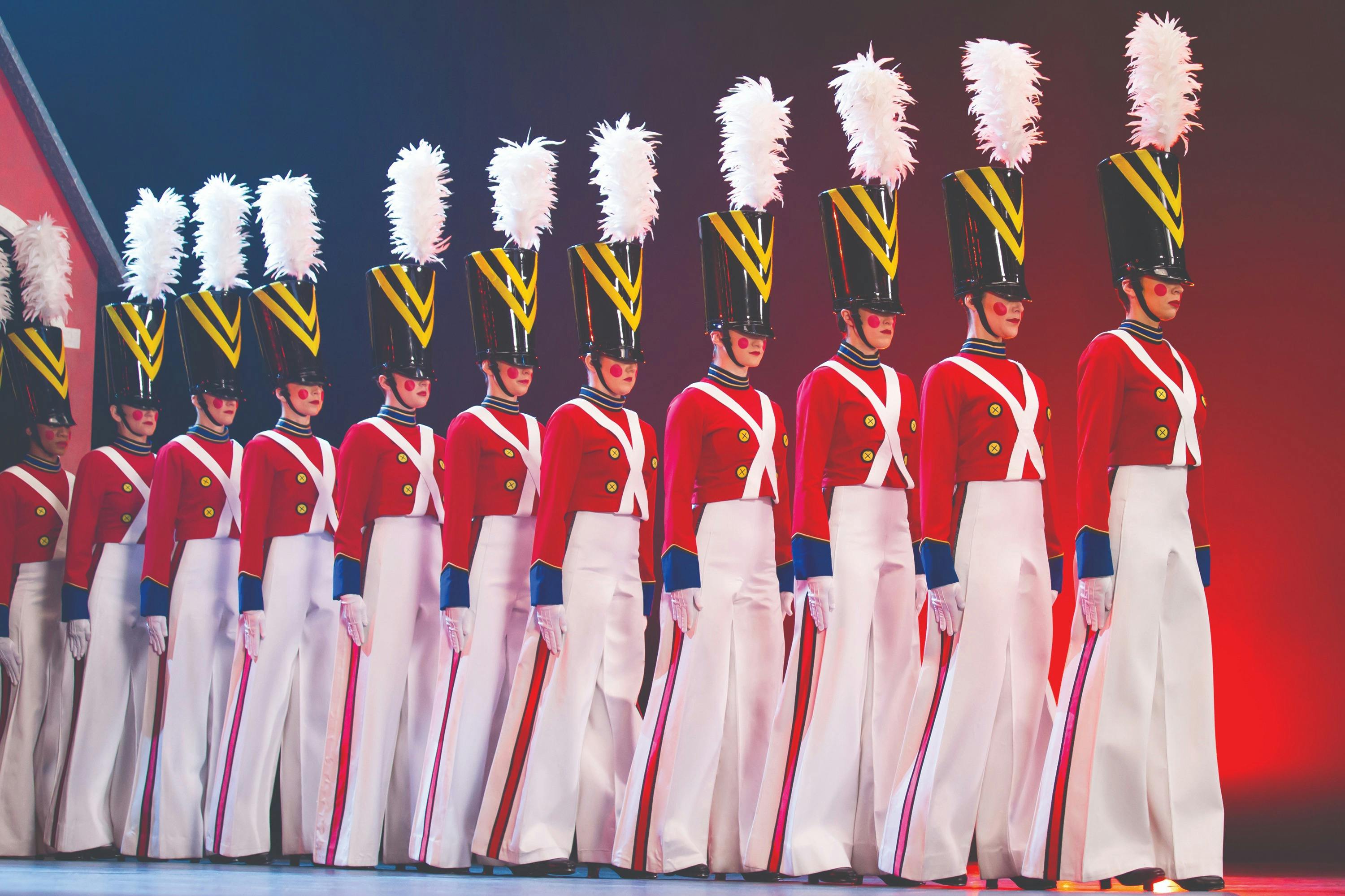 Um grupo de artistas vestidos de soldadinhos de chumbo, com uniformes vermelhos e brancos, chapéus altos e plumas, em formação.
