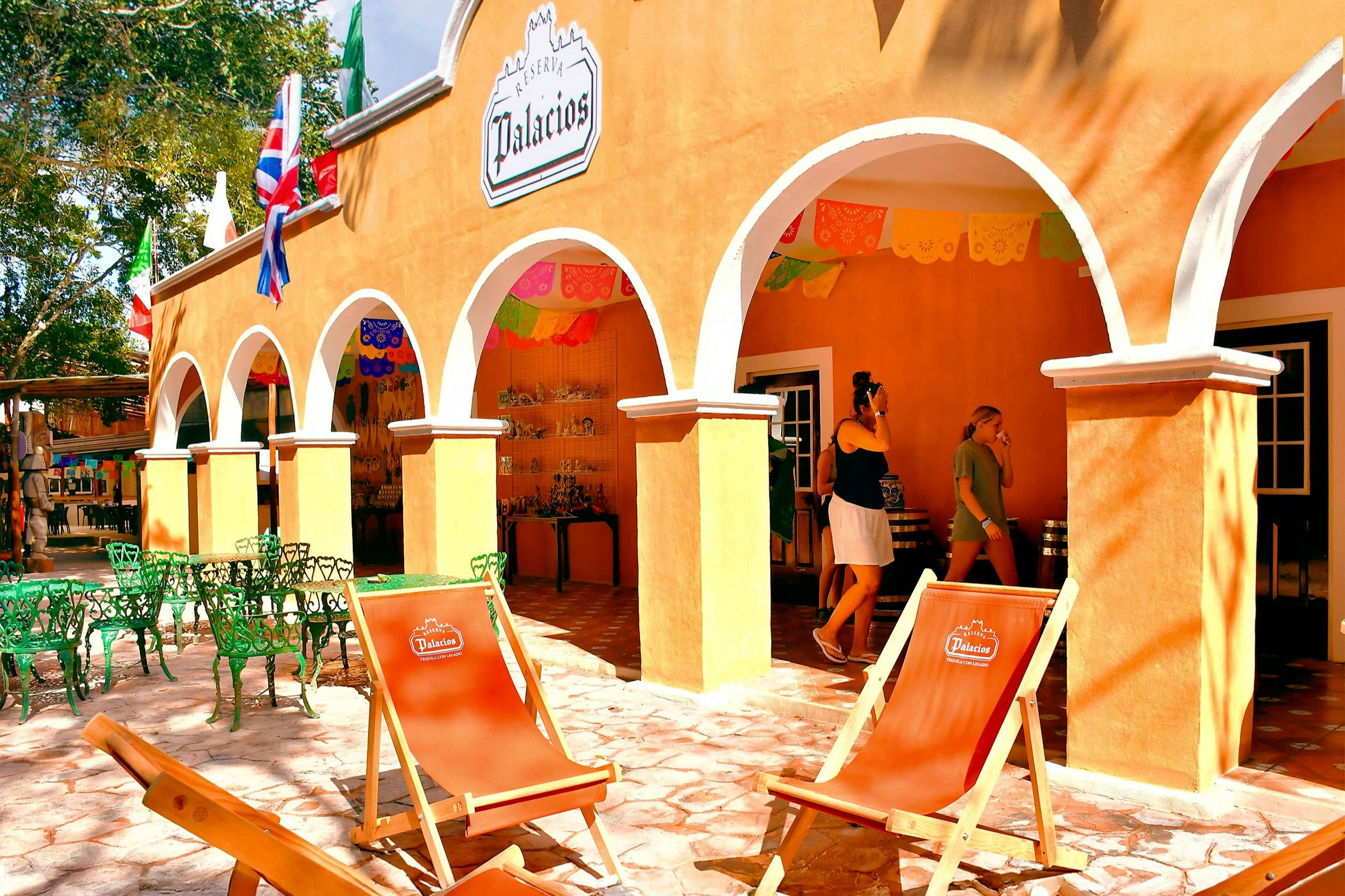 Outdoor seating with orange deck chairs and a building with arched entrances, colorful banners, and a shop inside.