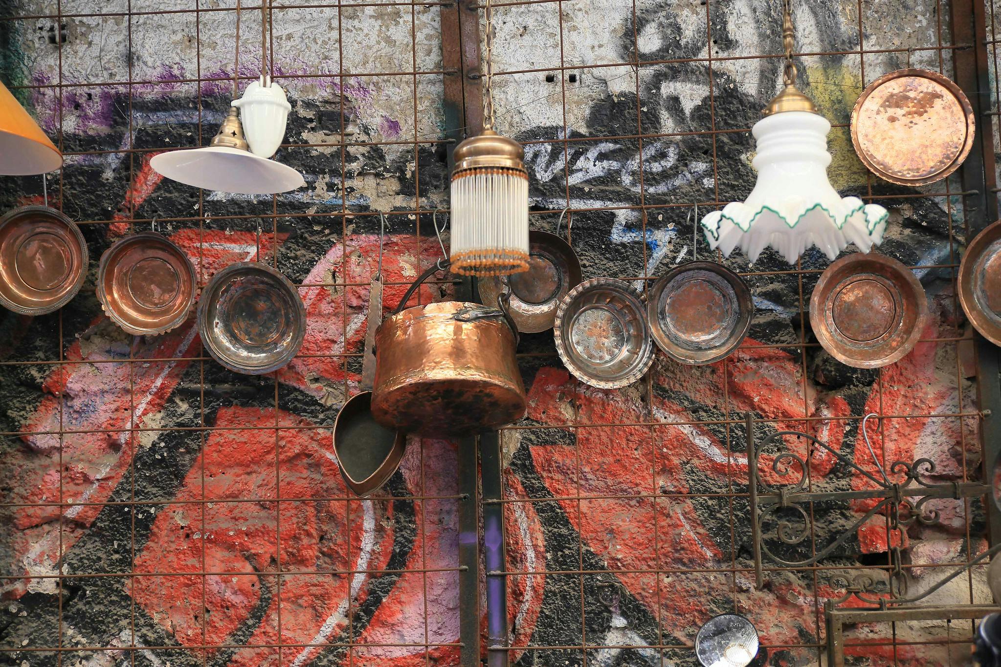 Hängande kopparkrukor och tallrikar, tillsammans med vintage-lampor, visas mot en färgglad graffitibakgrund på ett metallgaller.