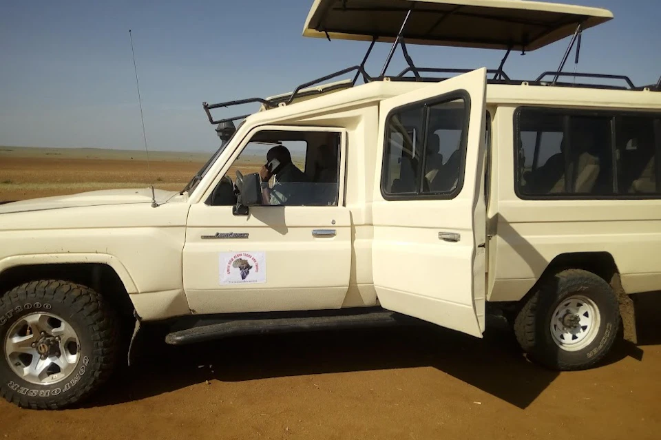 A white safari vehicle with an open roof and a logo on the side. A person is seated in the driver's seat on a phone.