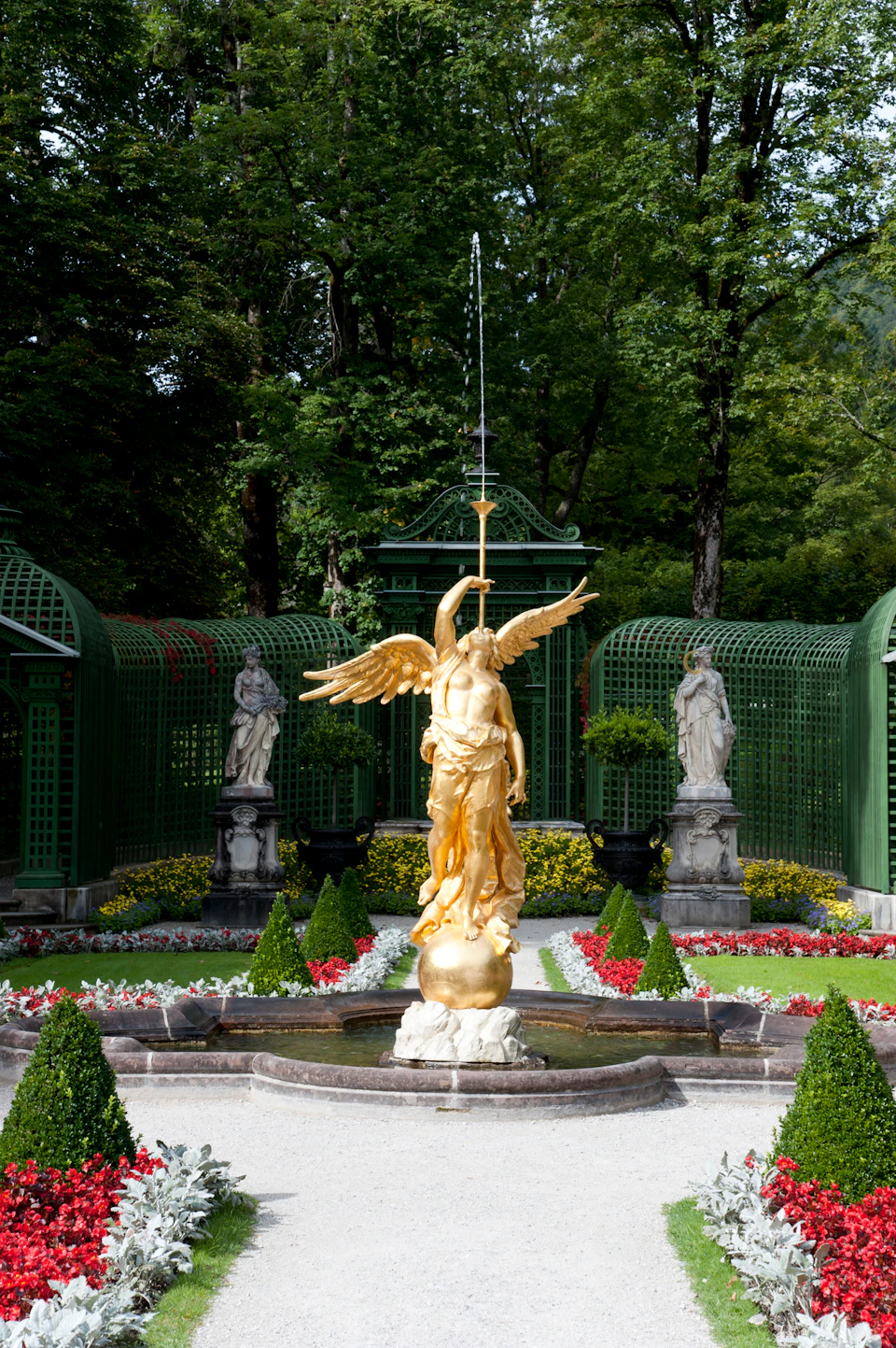 A golden statue of an angel holding a spear stands in a landscaped garden, surrounded by greenery and decorative sculptures.