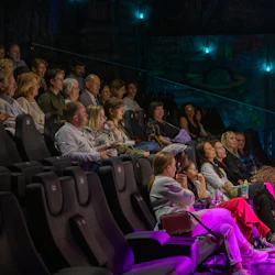 Audience seated in a dimly lit theater, watching a performance or presentation, with colorful lighting illuminating the scene.
