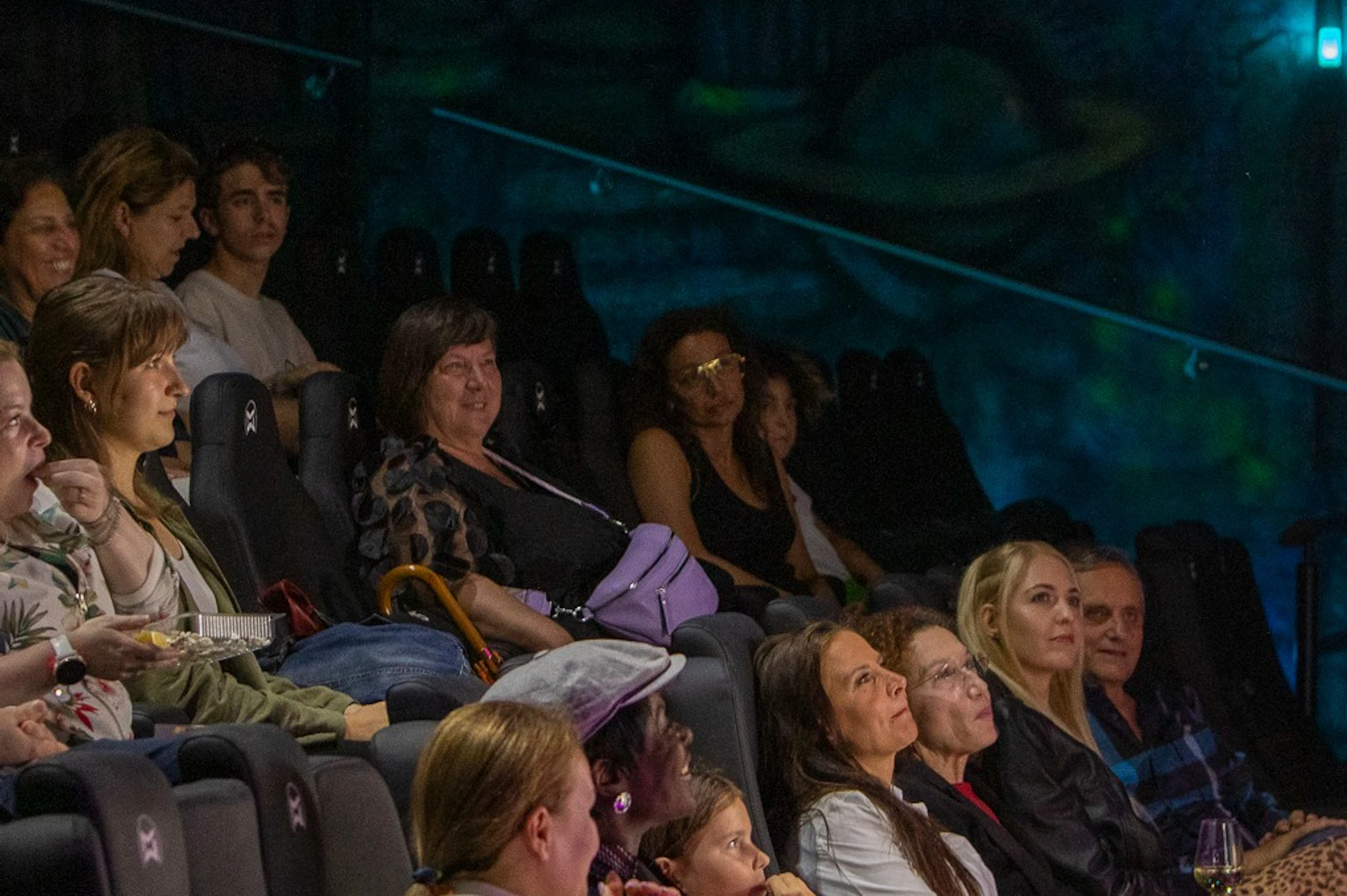 Audience seated in a dimly lit theater, watching a performance or presentation, with colorful lighting illuminating the scene.