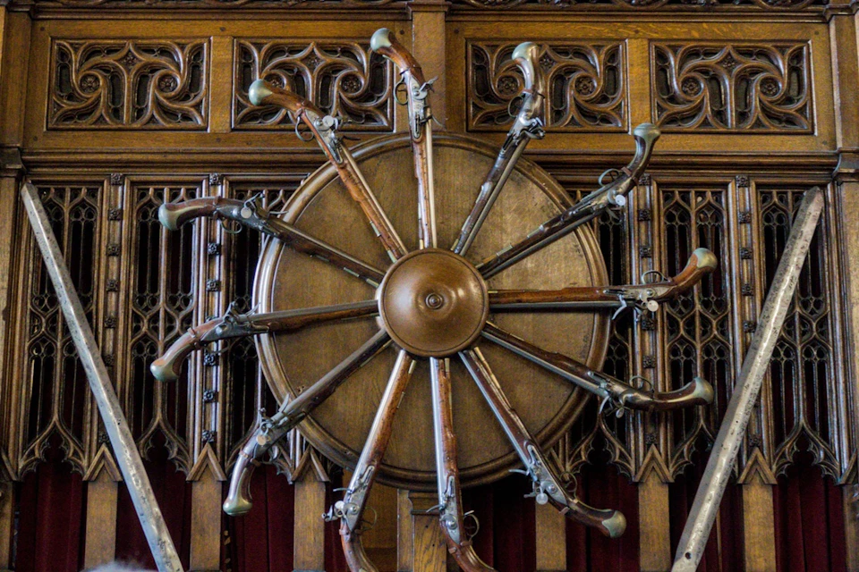 A circular wooden display featuring pistols arranged radially, set against ornate wooden carvings on a wall.