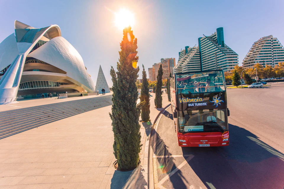 A red Valencia Bus Turistic drives near modern architecture under a bright sun, with tall trees and a clear sky in the background.