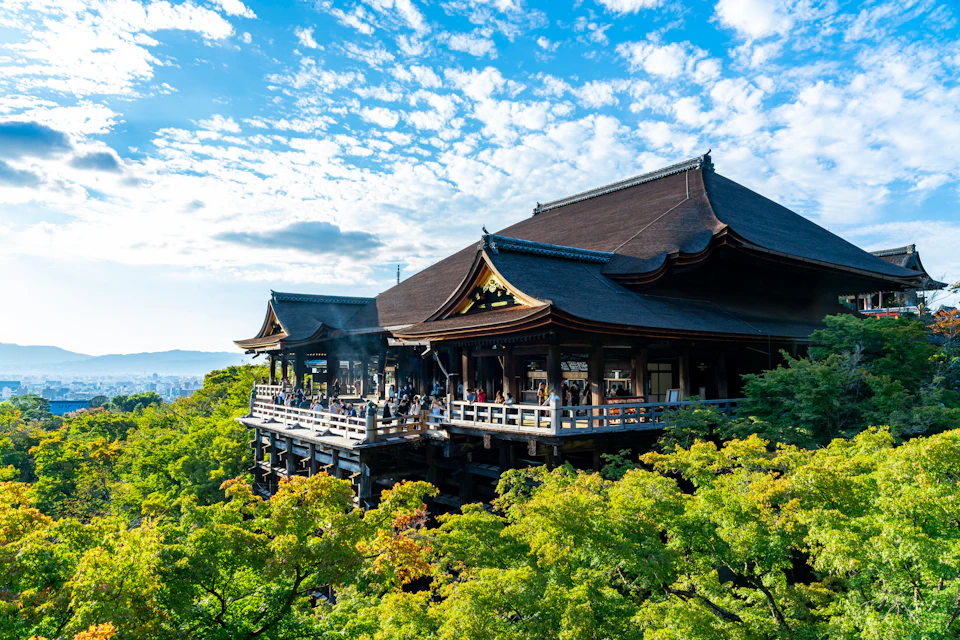 A traditional wooden temple with a large terrace surrounded by lush green trees under a partly cloudy blue sky.