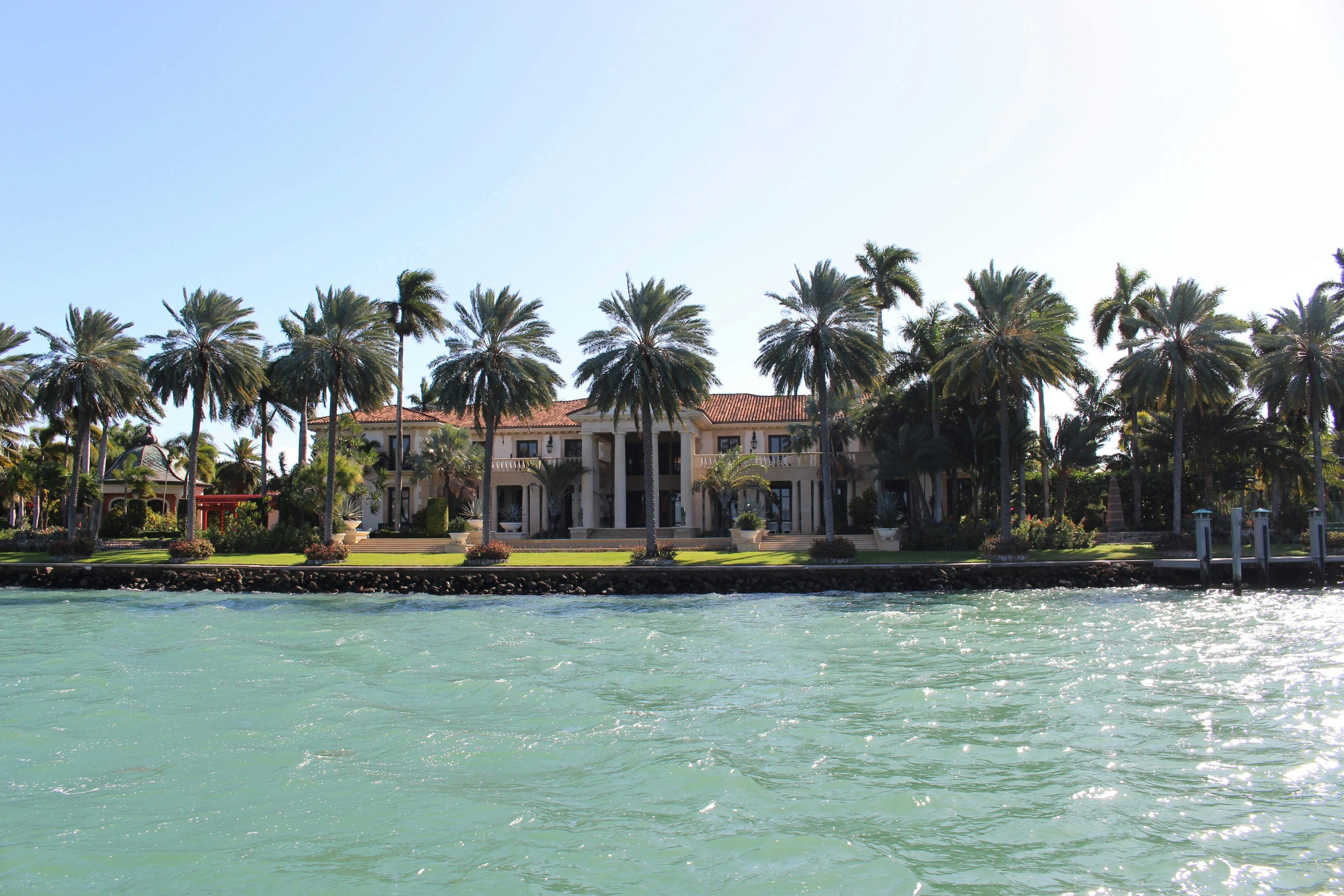 A waterfront mansion with a red tile roof, surrounded by palm trees, with a green lawn and water in the foreground.