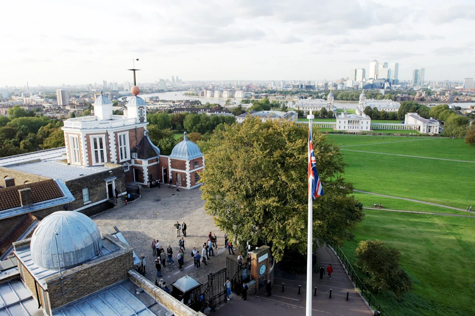 Overhead view of the Royal Observatory in Greenwich, with visitors, a large tree, and the Union Jack flag, overlooking London.