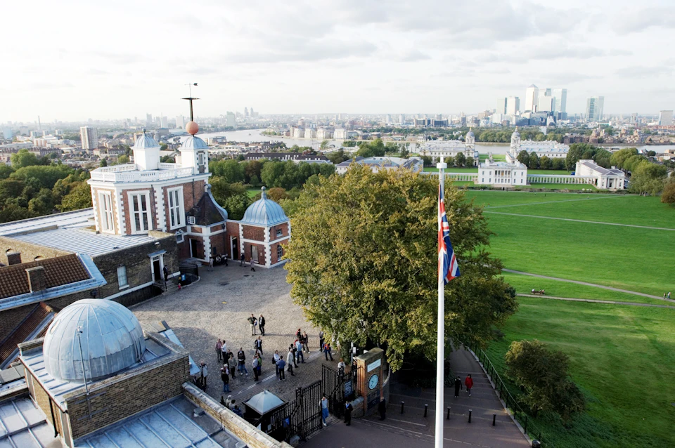 Overhead view of the Royal Observatory in Greenwich, with visitors, a large tree, and the Union Jack flag, overlooking London.