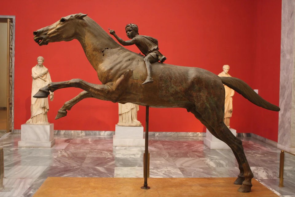 A bronze statue of a child riding a horse, displayed in a museum with a red wall and marble floor, surrounded by other statues.