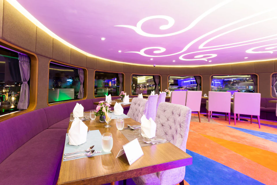 Upscale restaurant interior with elegant table settings, plush chairs, and modern ceiling lighting, featuring large windows and colorful carpeting.