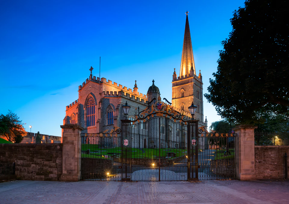 A historic, illuminated cathedral with a tall spire, seen at dusk behind ornate iron gates, surrounded by a paved area and trees.