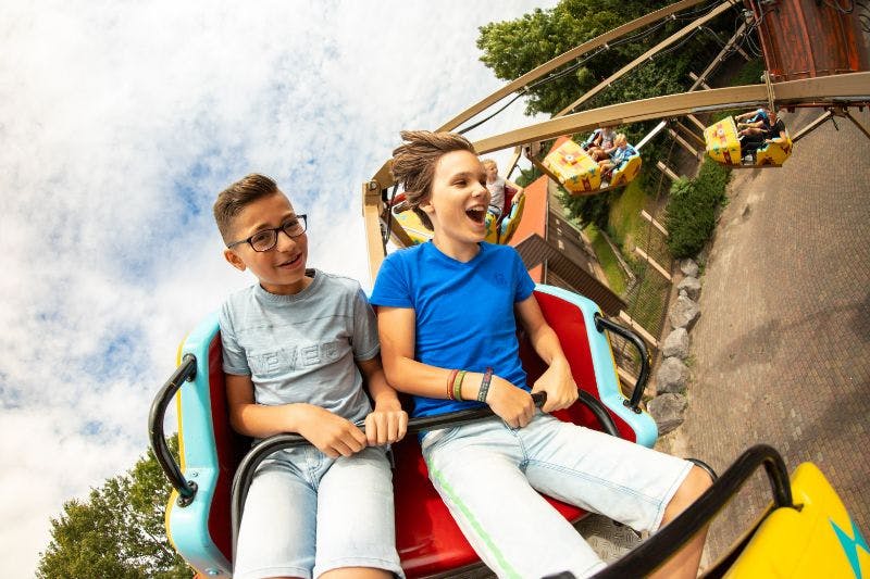 Two boys ride a roller coaster, smiling and laughing; the sky is partly cloudy with greenery in the background.