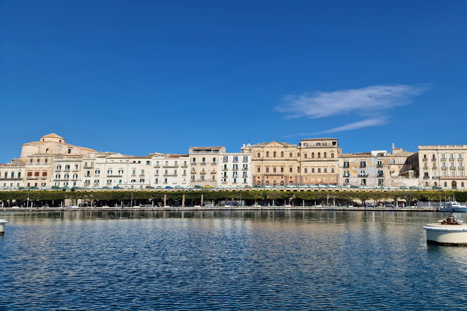 Row of historic buildings with balconies along a waterfront, under a clear blue sky with a few clouds.