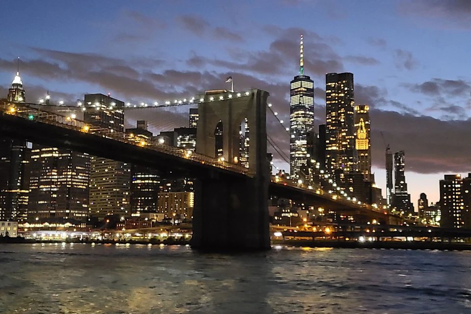 Brooklyn Bridge at night with illuminated Manhattan skyline in the background and reflections in the water below.