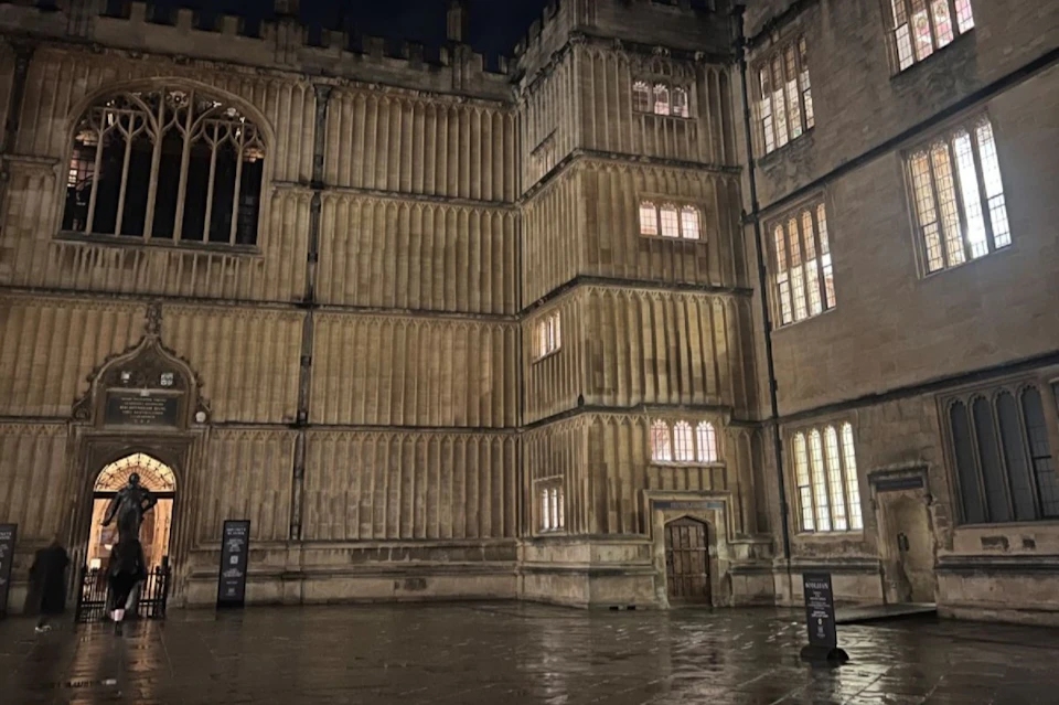 A historic stone building with illuminated windows at night, featuring intricate architectural details and a wet courtyard.