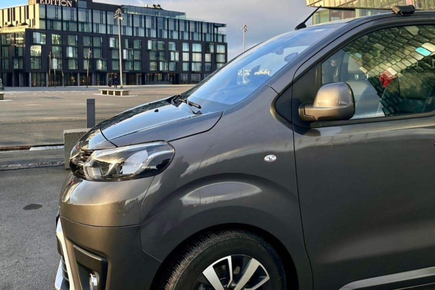 Modern, grey van parked on a street with a glass building in the background under a cloudy sky.