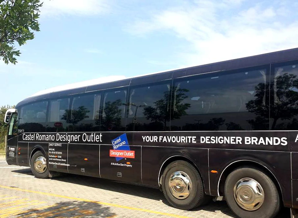 A black tour bus with "Castel Romano Designer Outlet" branding parked on a paved surface under a clear sky.