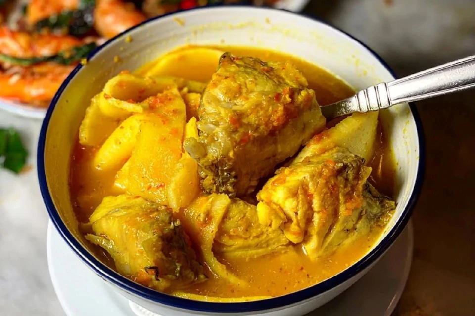 A bowl of fish stew with yellow broth, chunks of fish and potatoes, and a spoon visible inside the bowl.