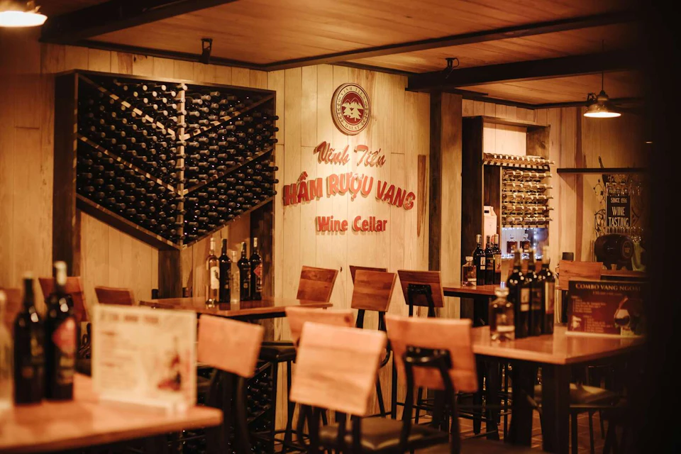 A cozy wine cellar with wooden decor, wine racks filled with bottles, and tables with chairs arranged for seating.