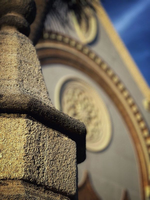 Nahaufnahme einer Steinsäule mit einem unscharfen verschnörkelten Zifferblatt an einem Gebäude im Hintergrund.