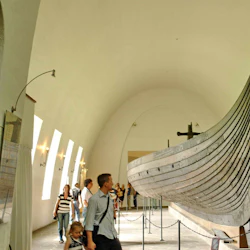 Besökare observerar ett stort, historiskt träskepp som visas i ett museum med välvda tak och riklig belysning.