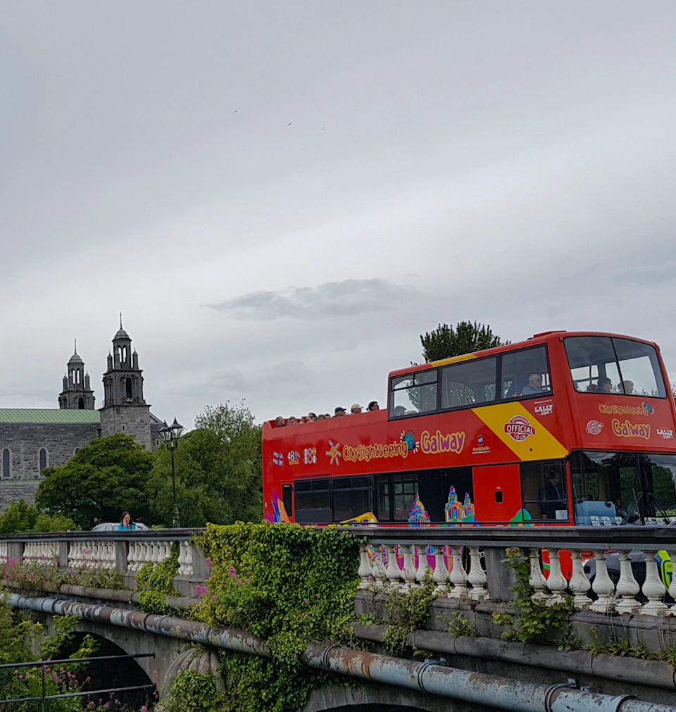 A red double-decker tour bus crossing a bridge with an old stone building with towers in the background.