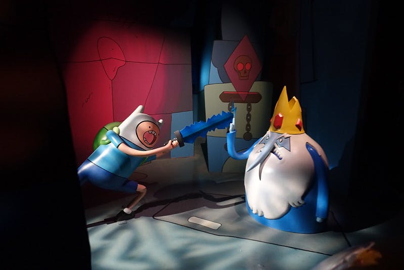 Ένα αγόρι κινουμένων σχεδίων με ένα σπαθί αντιμετωπίζει μια μεγάλη γενειοφόρο φιγούρα σε ένα στέμμα μέσα σε ένα αμυδρά φωτισμένο, πολύχρωμο δωμάτιο.