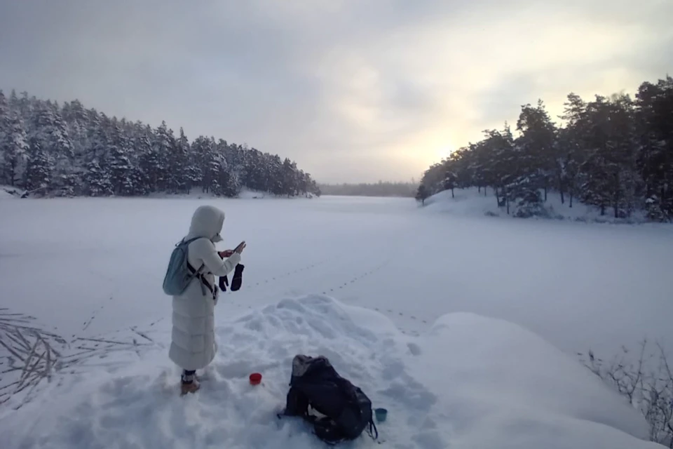 A person dressed in winter clothing stands on snow by a frozen lake, with trees in the background under a cloudy, bright sky.