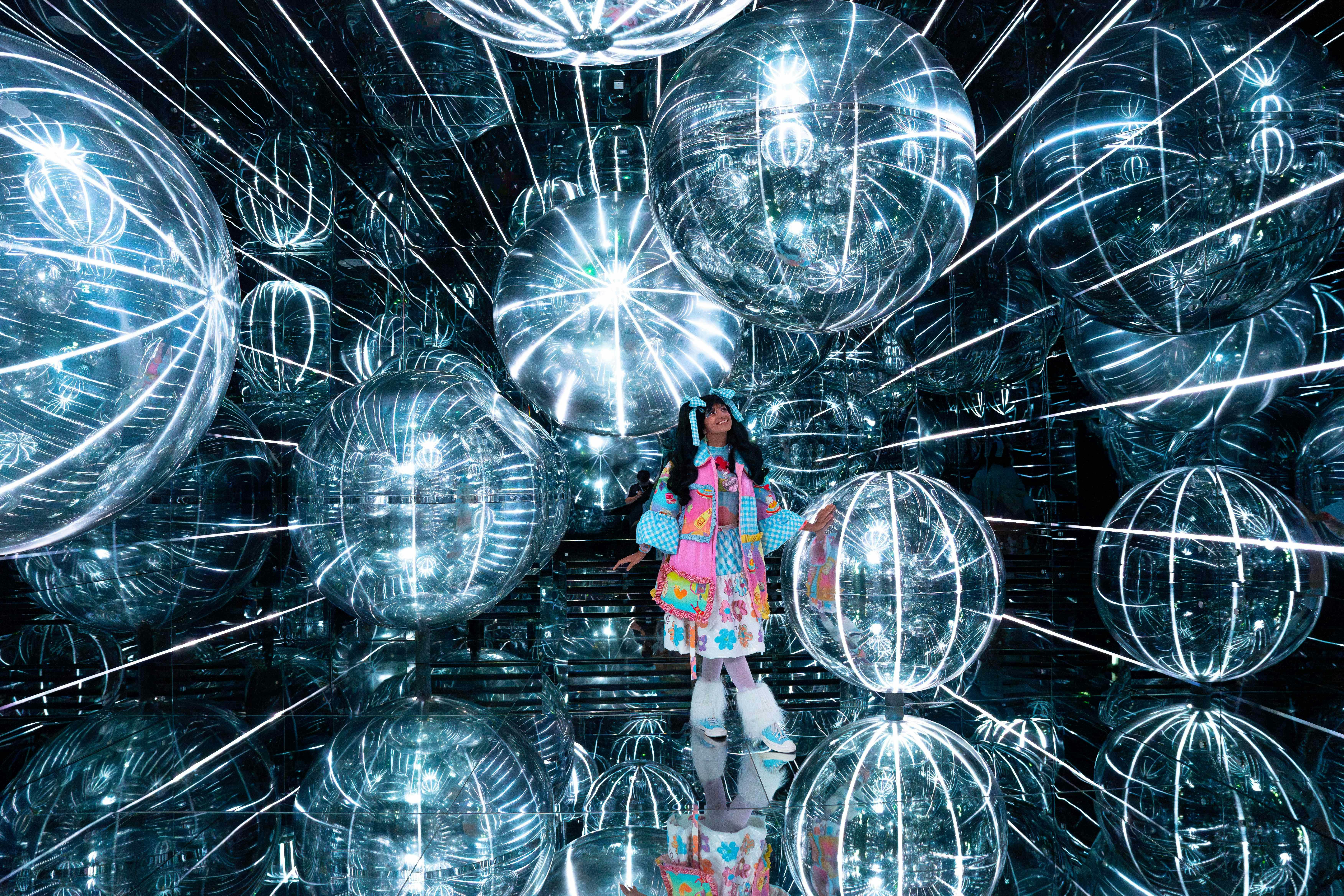 Osoba v barevném oblečení stojí v reflexní místnosti plné velkých osvětlených sférických objektů.