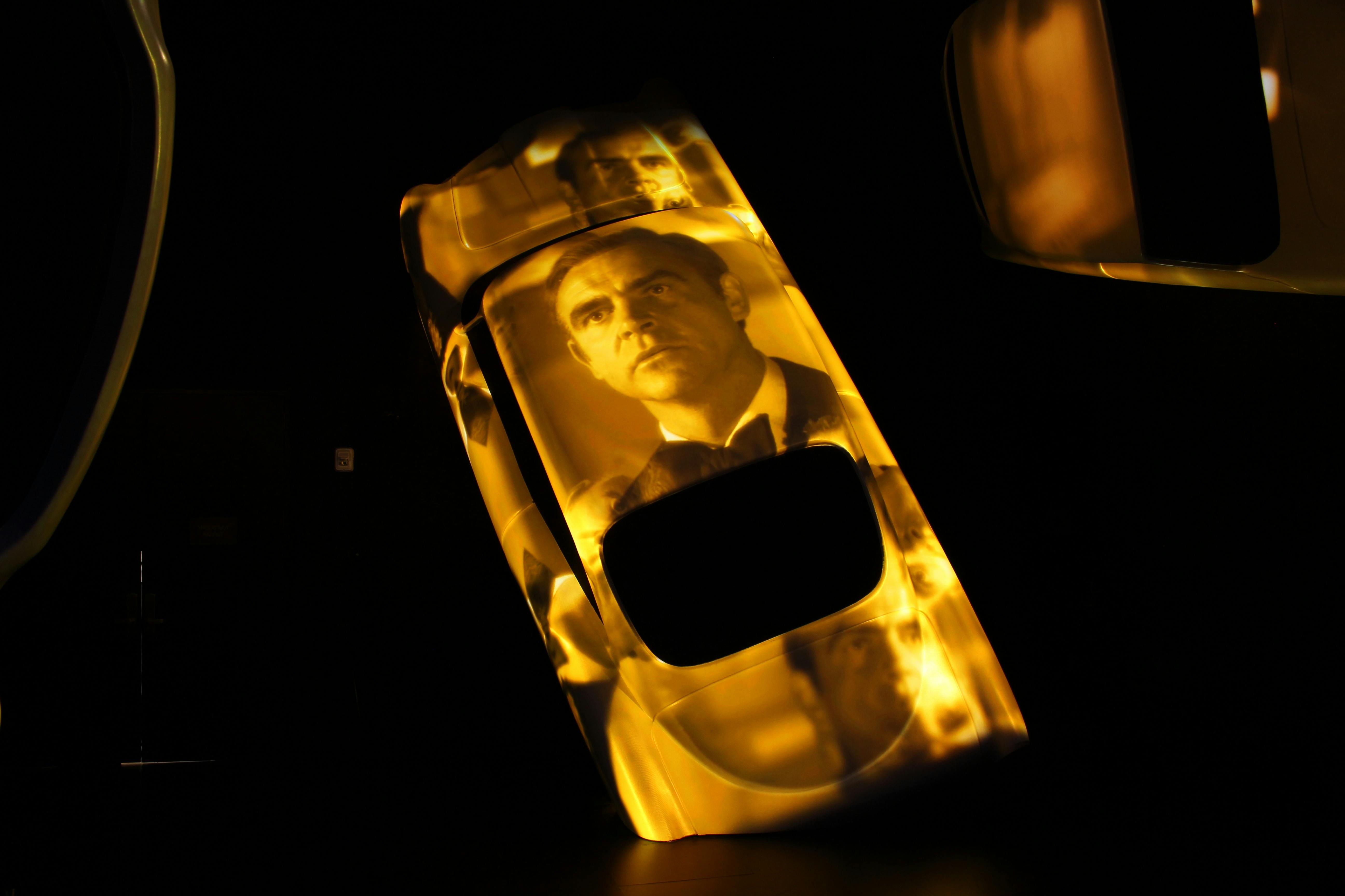 Ένα ανακλινόμενο αυτοκίνητο με κίτρινο φως και προβαλλόμενες εικόνες ενός άνδρα με κοστούμι σε σκούρο φόντο.