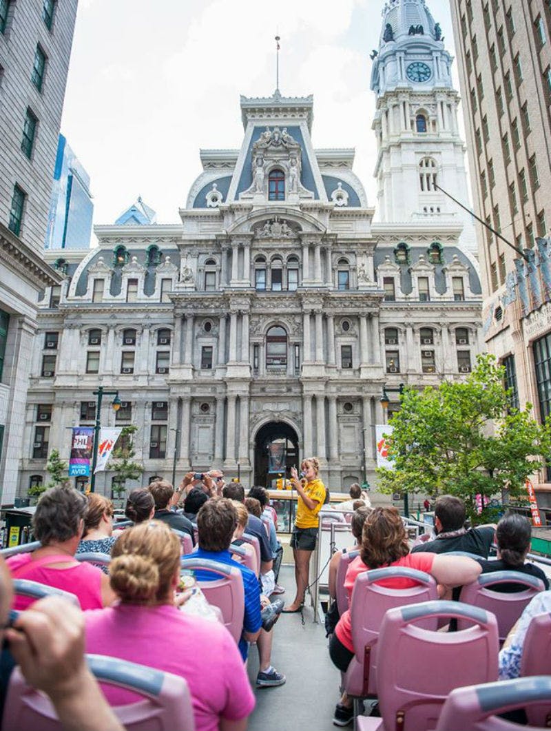 People seated on a tour bus, facing a guide standing, with a historic building and clock tower in the background.