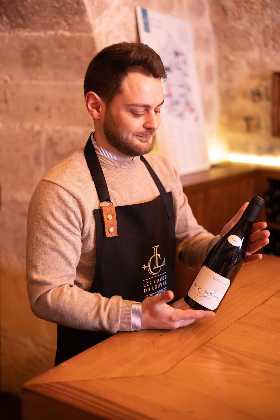 검은색 앞치마를 두른 남성이 돌담과 흐릿한 포스터를 배경으로 따뜻하게 빛나는 방에서 와인병을 들고 있습니다.