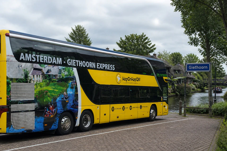 A yellow and black double-decker bus labeled "Amsterdam - Giethoorn Express" parked near a canal under a cloudy sky.