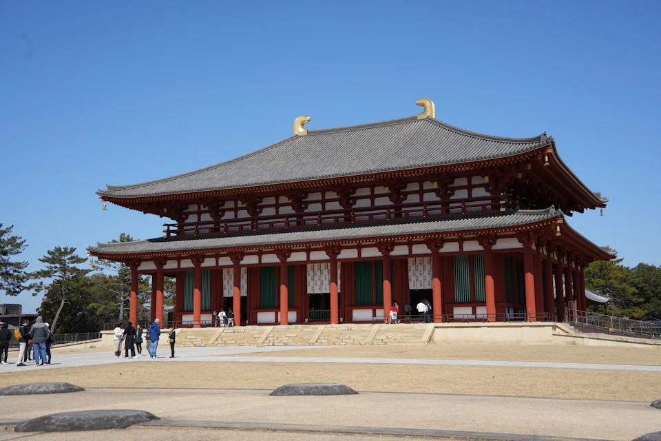 A traditional Japanese temple with red columns and a tiled roof under a clear blue sky, people walking nearby.