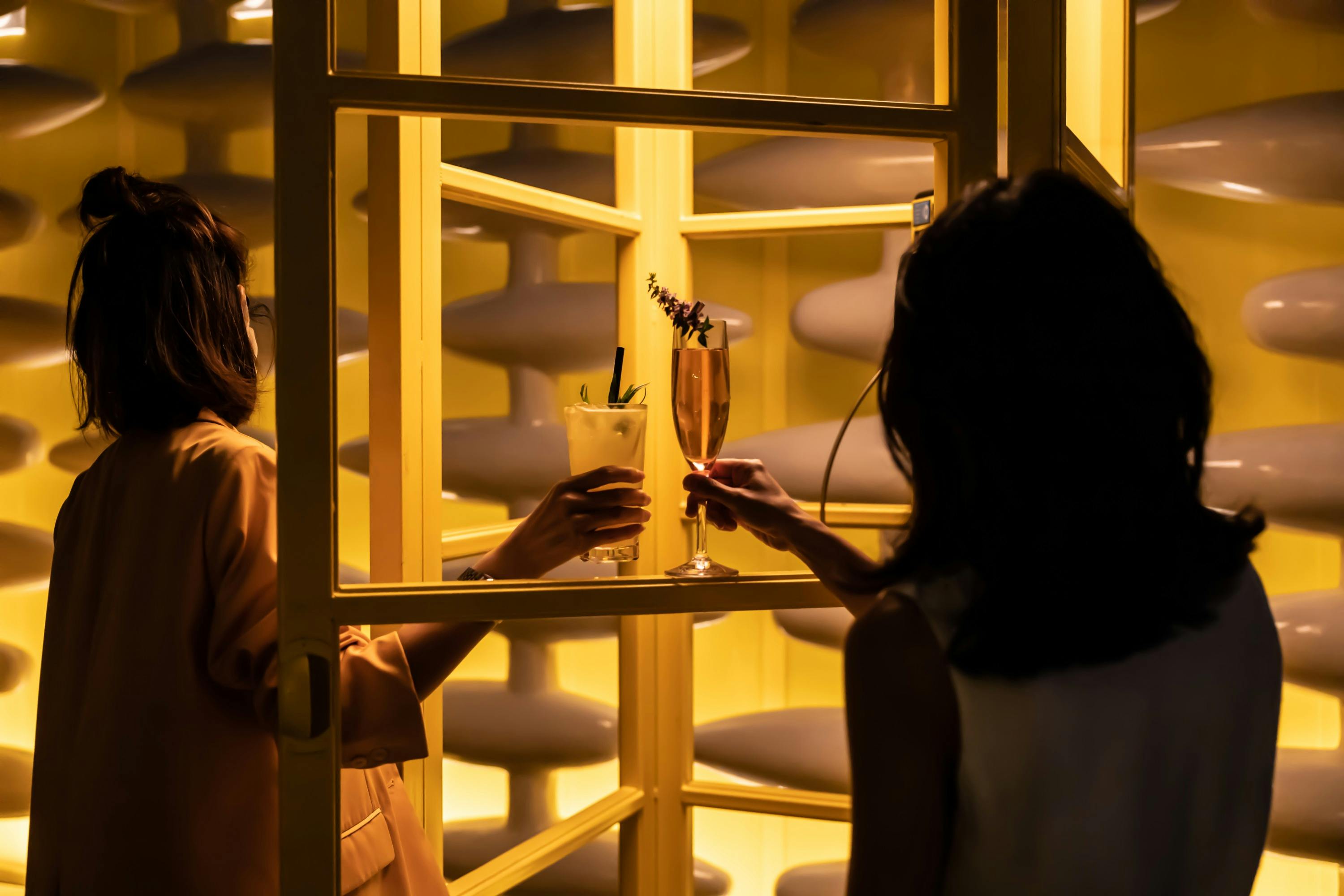 Two people clinking glasses, one with a cocktail and the other with a champagne flute, near yellow illuminated shelves.