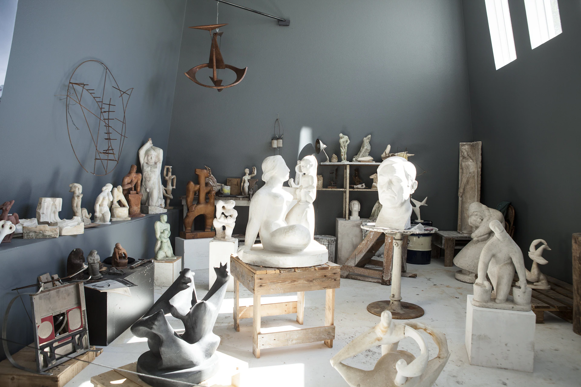 A studio filled with various sculptures, including human figures, abstract shapes, and busts, arranged on tables and shelves.