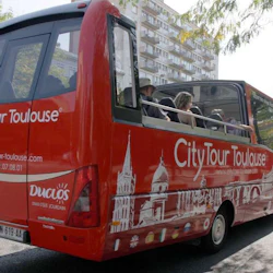 Red CityTour bus with passengers, open-top roof, and Toulouse landmarks depicted on the side driving near a residential building.