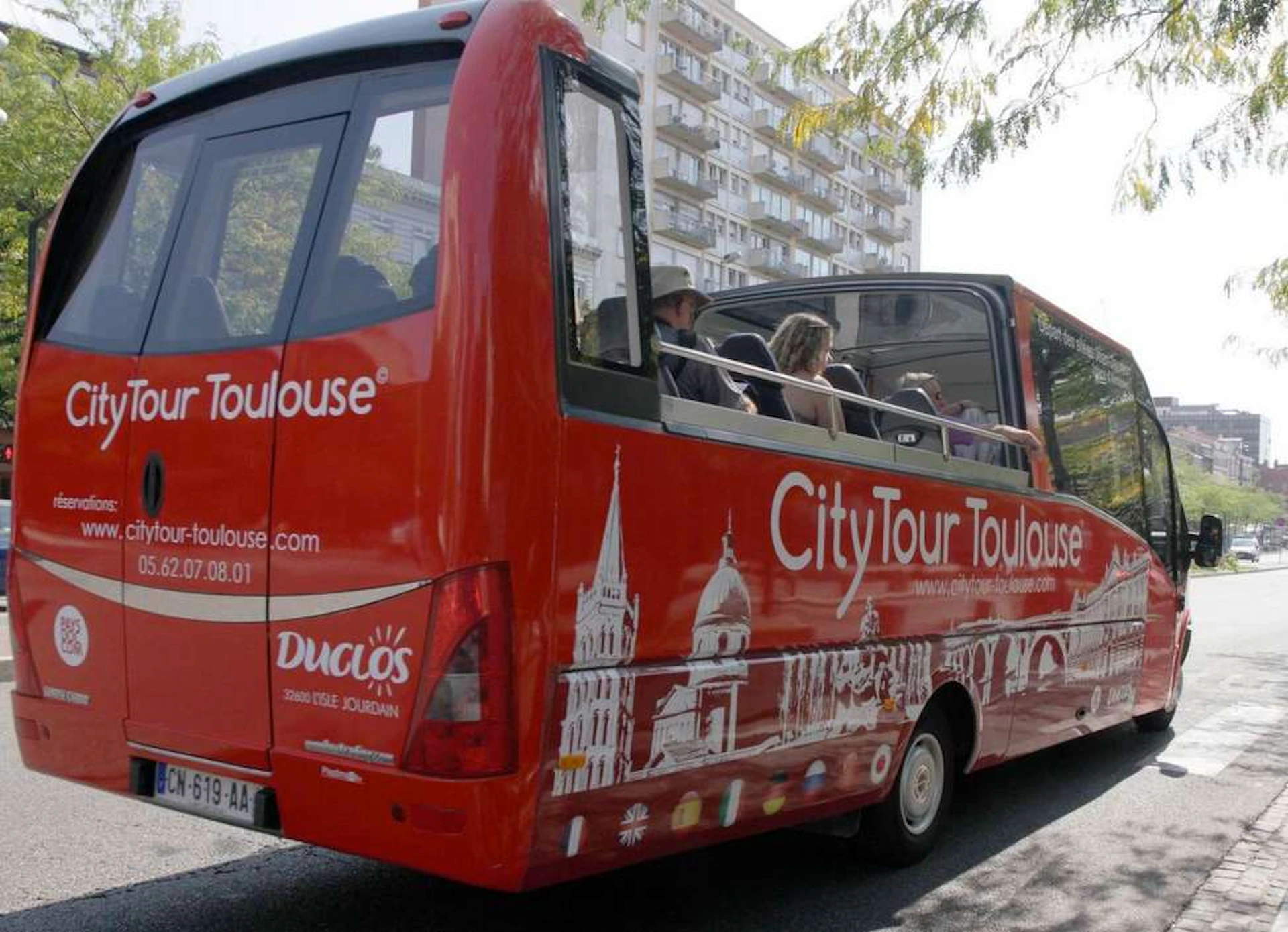 Red CityTour bus with passengers, open-top roof, and Toulouse landmarks depicted on the side driving near a residential building.