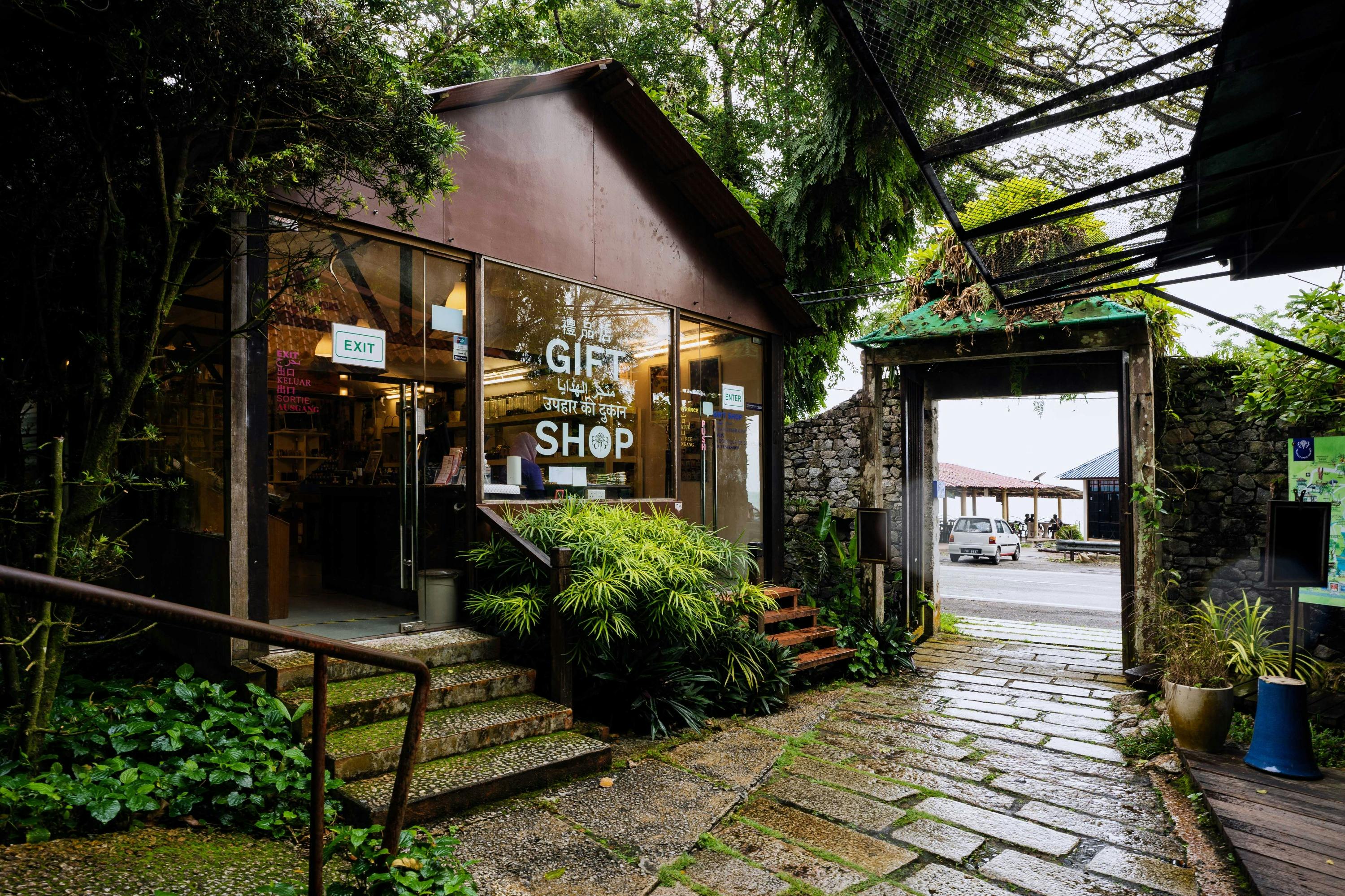 A gift shop with glass walls surrounded by greenery, near a cobblestone pathway and an open gate leading outside.