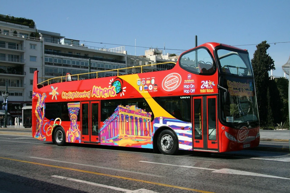 A double-decker sightseeing bus in Athens with colorful artwork of local landmarks, parked on a city street.