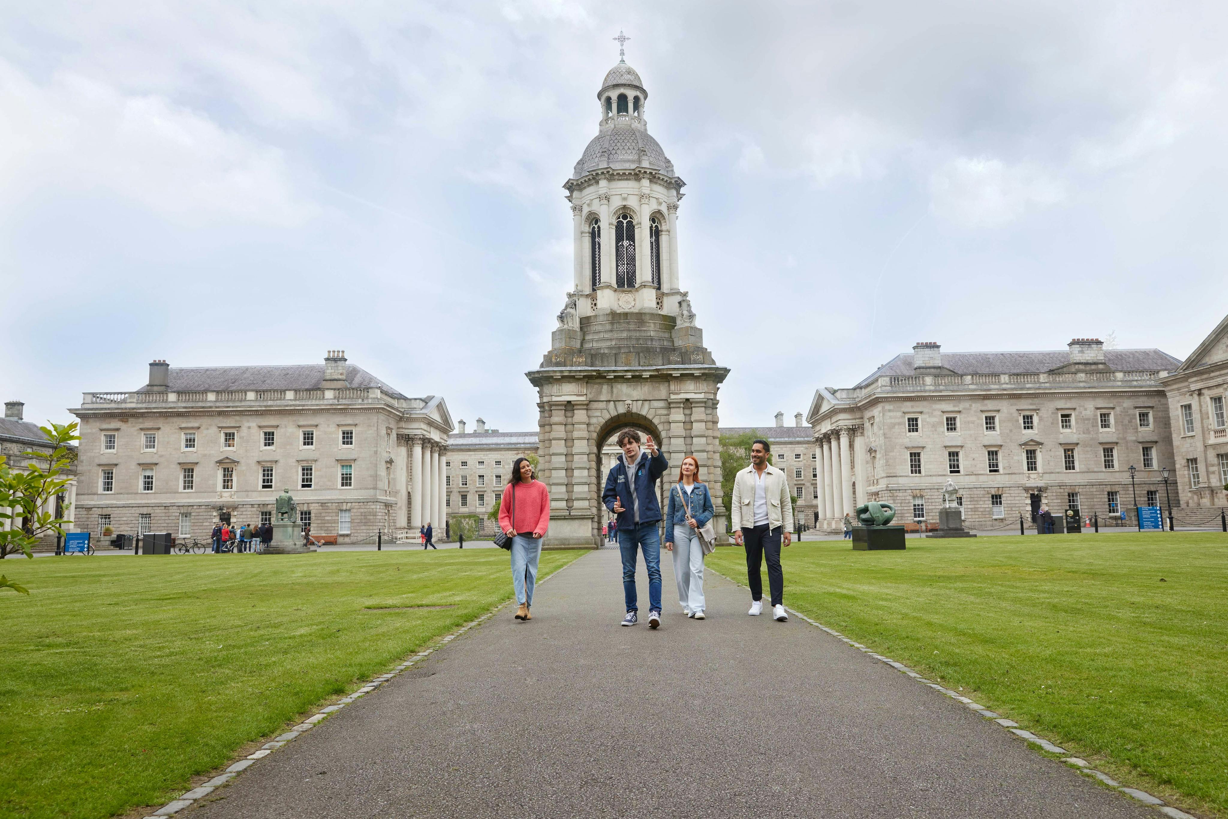 Four people walk on a pathway in front of a historic building with a large tower, flanked by other similar buildings.